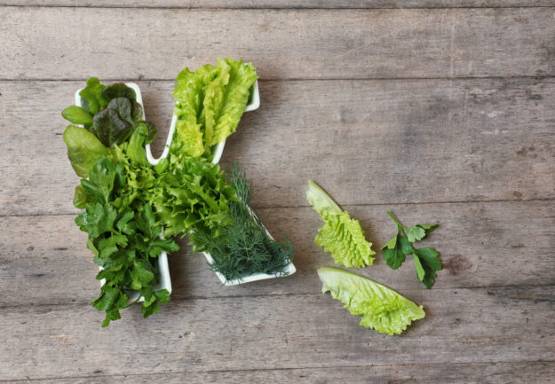 Vitamin K in food concept. Plate in the shape of the letter K with different fresh leafy green vegetables,  lettuce, herbs on wooden background. Flat lay or top view.